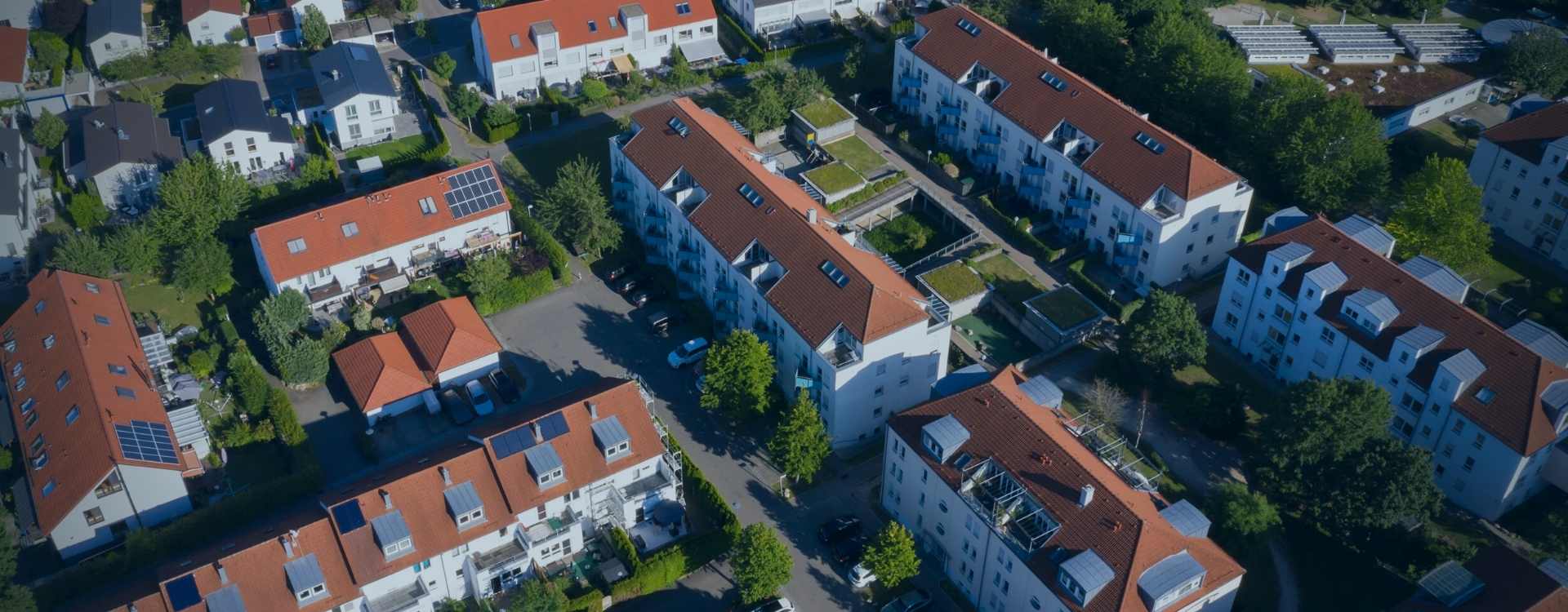 A residential area was photographed from a bird's eye's view, with small streets and green areas between the houses. In the middle and on the right, you can see several 5-story apartment buildings with balconies and parking lots in the front. Smaller multifamily houses can be seen on the left.