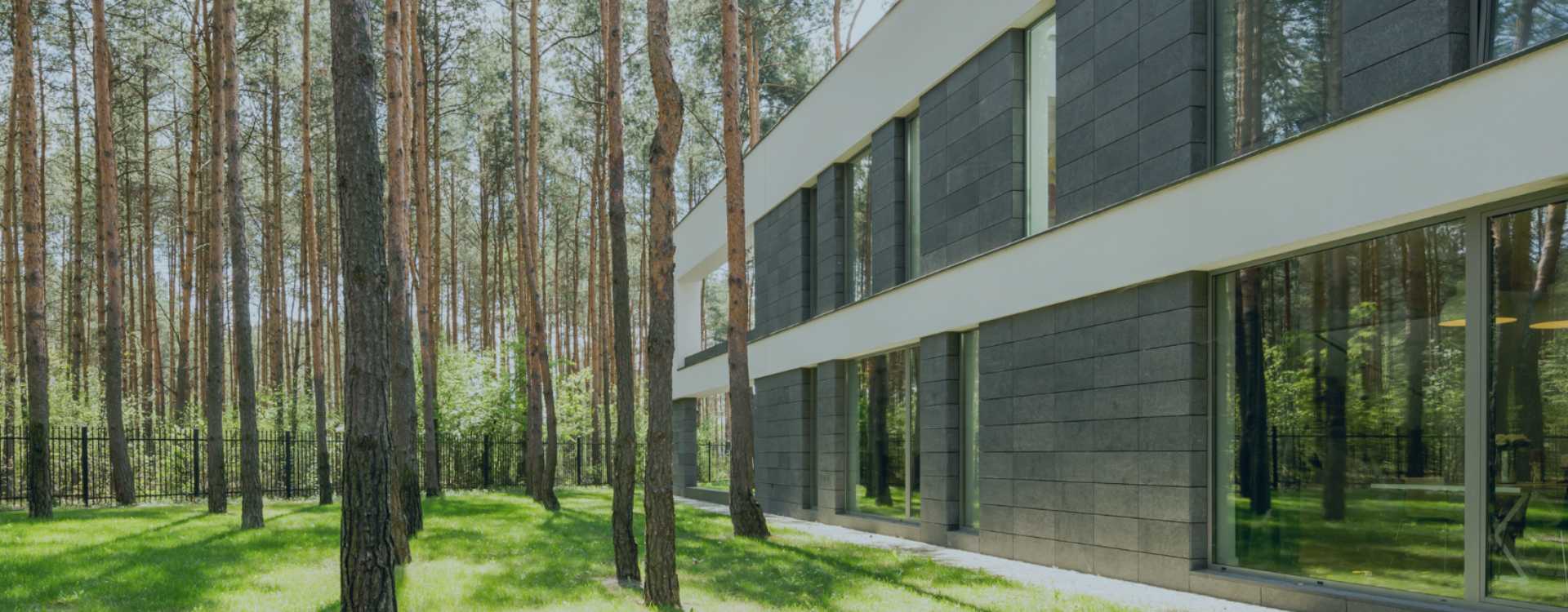 On the right side of the picture, a modern house with large windows and a fenced garden with large trees to its left can be seen standing in a forest.